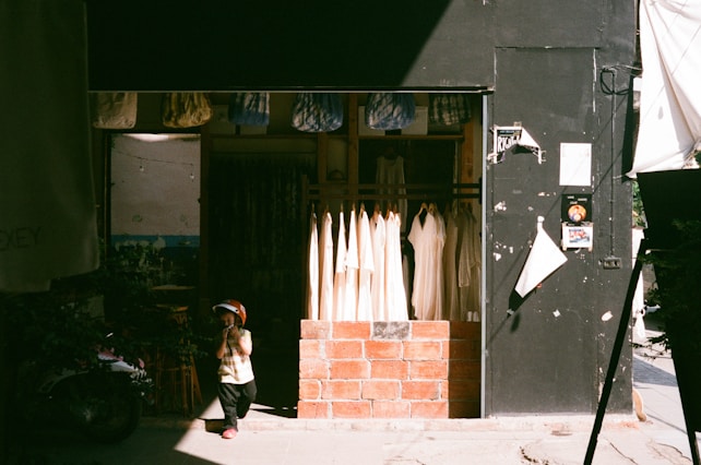 A small child in a helmet stands in front of a clothing shop. The shop features hanging white garments, and its entrance is framed with brick. The child's shadow is cast on the pavement, and the shop is partly in shadow with sunlight streaming in.