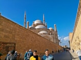 A large mosque with multiple domes and minarets is surrounded by tall stone walls under a clear blue sky. People are walking along a path beside the wall, with some individuals in the foreground looking towards the camera.