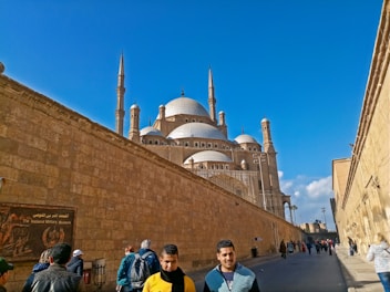 A large mosque with multiple domes and minarets is surrounded by tall stone walls under a clear blue sky. People are walking along a path beside the wall, with some individuals in the foreground looking towards the camera.