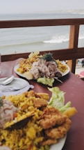 A table set with two plates of food, featuring yellow rice, fried seafood, and garnishes. The table is situated on a balcony overlooking a beach and ocean with waves visible in the background. The setting evokes a casual, seaside dining experience.