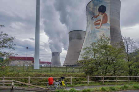 Several large cooling towers from an industrial facility dominate the landscape, with one tower featuring a mural of a child. Smoke or steam is seen billowing from the towers, creating a dramatic sky. In the foreground, there is a bicycle with bags parked along a road lined by a wooden fence. Trees add touches of green to the scene.