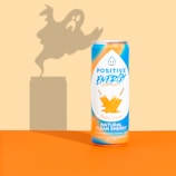 A can of Positive Energy drink is placed against a background with a ghostly shadow looming behind it. The can is decorated with vibrant orange and blue colors, featuring images of orange ice pops and text highlighting its natural clean energy content.