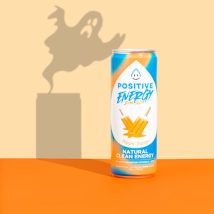 A can of Positive Energy drink is placed against a background with a ghostly shadow looming behind it. The can is decorated with vibrant orange and blue colors, featuring images of orange ice pops and text highlighting its natural clean energy content.