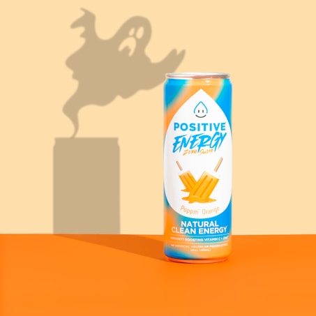 A can of Positive Energy drink is placed against a background with a ghostly shadow looming behind it. The can is decorated with vibrant orange and blue colors, featuring images of orange ice pops and text highlighting its natural clean energy content.