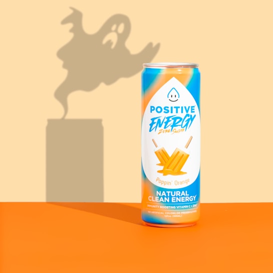 A can of Positive Energy drink is placed against a background with a ghostly shadow looming behind it. The can is decorated with vibrant orange and blue colors, featuring images of orange ice pops and text highlighting its natural clean energy content.