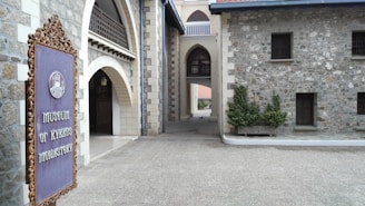 An architectural structure composed of gray stone brick walls with arched walkways and a detailed iron sign on the left. The sign reads 'Museum of Kykkos Monastery'. The structure appears to be historical, with small windows and decorative elements.