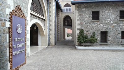 An architectural structure composed of gray stone brick walls with arched walkways and a detailed iron sign on the left. The sign reads 'Museum of Kykkos Monastery'. The structure appears to be historical, with small windows and decorative elements.