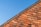 A section of a red brick roof is captured against a clear blue sky. The roof tiles are arranged in a neat, overlapping pattern, creating a textured and orderly geometric design.