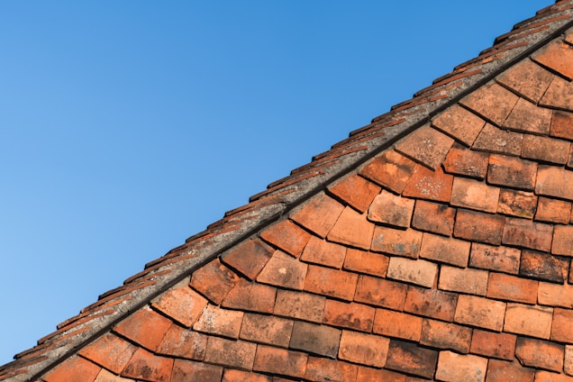 A section of a red brick roof is captured against a clear blue sky. The roof tiles are arranged in a neat, overlapping pattern, creating a textured and orderly geometric design.