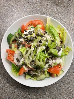 A fresh salad served in a white bowl, featuring crisp lettuce, sliced tomatoes, and cucumbers. The salad is topped with a creamy dressing and sprinkled with a mix of seeds, including pumpkin and flax seeds.