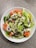 A fresh salad served in a white bowl, featuring crisp lettuce, sliced tomatoes, and cucumbers. The salad is topped with a creamy dressing and sprinkled with a mix of seeds, including pumpkin and flax seeds.