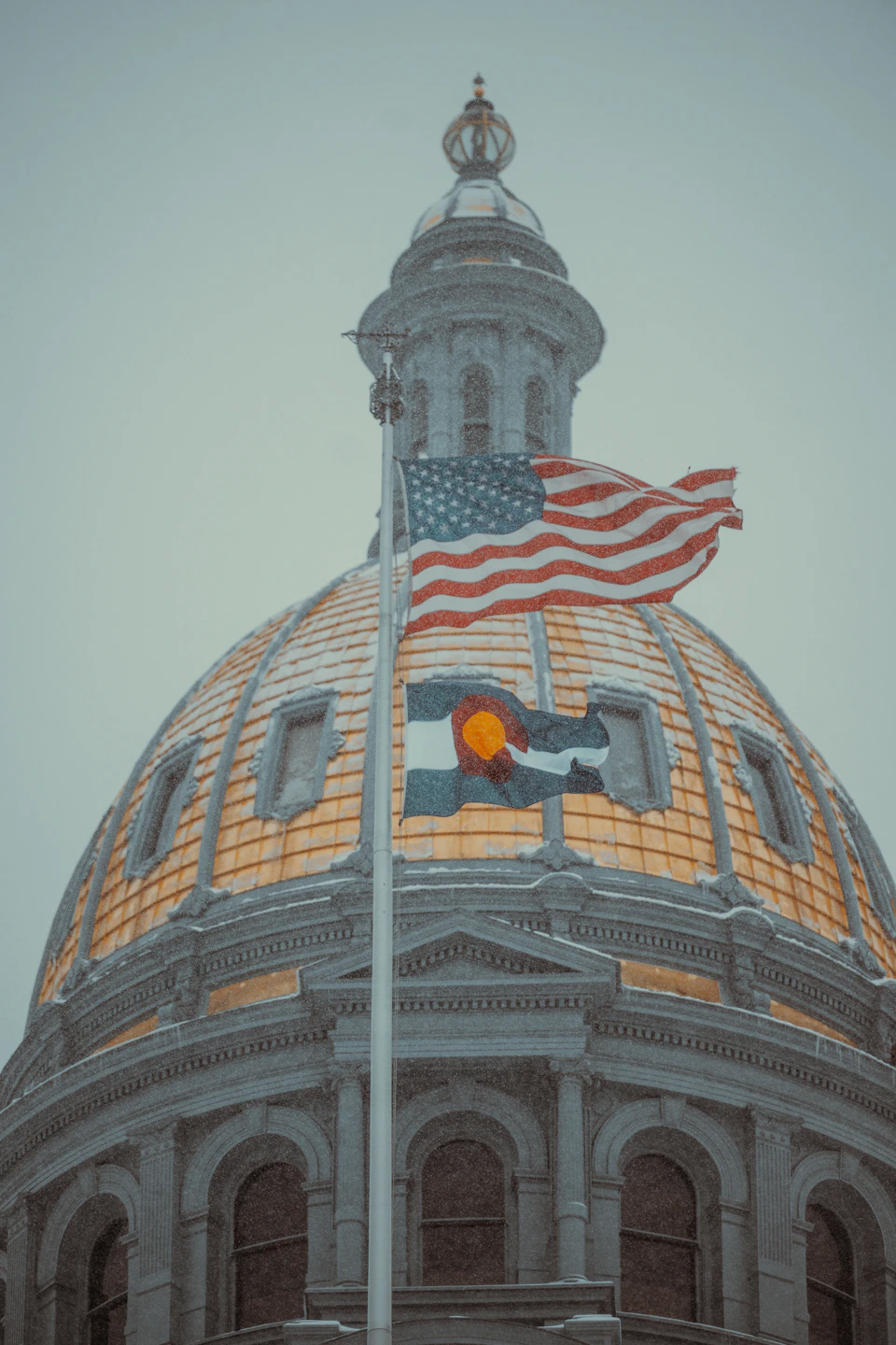 The image shows a majestic, large dome-shaped building with a gold and white facade that gleams under the light. The building is adorned with two American flags fluttering atop it - one on each side. The perspective of the photo is from below, looking up at the building, giving it an imposing presence against the sky.

The sky above the building is overcast, casting a soft light over the scene and creating a sense of depth in the image.