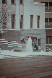 A small snowplow vehicle is actively clearing snow from a sidewalk in an urban setting. The snowfall creates a wintry atmosphere, with snow accumulating on the surrounding buildings and bare tree branches. The architecture features a modern design, and the street appears wet from the melted snow.