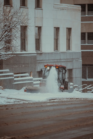 A small snowplow vehicle is actively clearing snow from a sidewalk in an urban setting. The snowfall creates a wintry atmosphere, with snow accumulating on the surrounding buildings and bare tree branches. The architecture features a modern design, and the street appears wet from the melted snow.