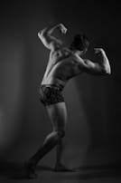 A muscular person poses in a classic bodybuilder stance, showcasing their back and arm muscles. The subject is wearing dark underwear and stands barefoot against a plain background. The lighting casts dramatic shadows, emphasizing muscle definition.