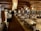 A spacious, rustic dining room with wooden interior featuring log walls and wooden beams along the ceiling. The room is filled with neatly arranged tables and chairs, set with glasses and napkins, ready for guests. Decorative light fixtures hang from the ceiling, and a large stone fireplace is located at one end of the room. There is a service area on one side with plates stacked neatly.
