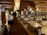 A spacious, rustic dining room with wooden interior featuring log walls and wooden beams along the ceiling. The room is filled with neatly arranged tables and chairs, set with glasses and napkins, ready for guests. Decorative light fixtures hang from the ceiling, and a large stone fireplace is located at one end of the room. There is a service area on one side with plates stacked neatly.