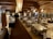 A spacious, rustic dining room with wooden interior featuring log walls and wooden beams along the ceiling. The room is filled with neatly arranged tables and chairs, set with glasses and napkins, ready for guests. Decorative light fixtures hang from the ceiling, and a large stone fireplace is located at one end of the room. There is a service area on one side with plates stacked neatly.