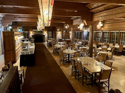 A spacious, rustic dining room with wooden interior featuring log walls and wooden beams along the ceiling. The room is filled with neatly arranged tables and chairs, set with glasses and napkins, ready for guests. Decorative light fixtures hang from the ceiling, and a large stone fireplace is located at one end of the room. There is a service area on one side with plates stacked neatly.