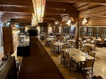 A spacious, rustic dining room with wooden interior featuring log walls and wooden beams along the ceiling. The room is filled with neatly arranged tables and chairs, set with glasses and napkins, ready for guests. Decorative light fixtures hang from the ceiling, and a large stone fireplace is located at one end of the room. There is a service area on one side with plates stacked neatly.