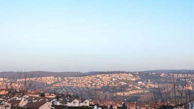 A scenic view of a sprawling residential area nestled among rolling hills. The landscape is dotted with numerous houses with red-tiled roofs, extending towards the horizon. Bare trees in the foreground suggest a winter or early spring setting. The sky is a clear blue, providing a serene backdrop.