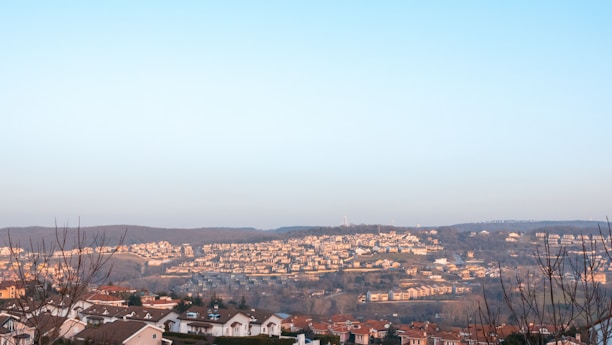 A scenic view of a sprawling residential area nestled among rolling hills. The landscape is dotted with numerous houses with red-tiled roofs, extending towards the horizon. Bare trees in the foreground suggest a winter or early spring setting. The sky is a clear blue, providing a serene backdrop.