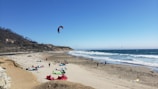 A sandy beach with several colorful kites and kite surfers. The ocean waves can be seen crashing onto the shore while cliffs line the background. People are either setting up or engaged in kitesurfing activities.