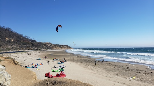 A sandy beach with several colorful kites and kite surfers. The ocean waves can be seen crashing onto the shore while cliffs line the background. People are either setting up or engaged in kitesurfing activities.