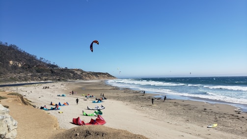 A sandy beach with several colorful kites and kite surfers. The ocean waves can be seen crashing onto the shore while cliffs line the background. People are either setting up or engaged in kitesurfing activities.