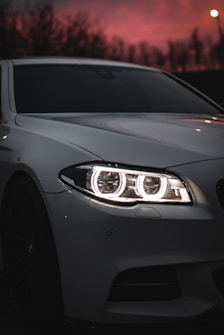 A sleek white car with bright, circular headlights is parked under a dramatic, reddish-pink sunset sky. The image focuses on the front section of the car, highlighting the design and intricate detailing of the headlights. In the background, dark silhouettes of trees can be faintly seen, adding depth to the composition.