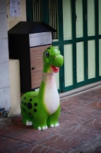 A colorful green dinosaur figure with a friendly expression stands on a brick sidewalk. It has a large smile and black spots on its body. Behind it, there is a green and white paneled door beside a black and wooden mailbox.