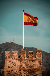 A weathered stone fortress with a Spanish flag fluttering high on a pole. The fortress features ancient brickwork, set against a mountainous background with communication towers visible on the peak. The sky is overcast yet bright, adding a sense of grandeur to the setting.