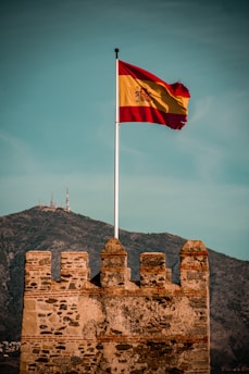A weathered stone fortress with a Spanish flag fluttering high on a pole. The fortress features ancient brickwork, set against a mountainous background with communication towers visible on the peak. The sky is overcast yet bright, adding a sense of grandeur to the setting.