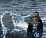 A person stands in front of a light projection that creates abstract patterns and lines, casting shadows on their body. The patterns are predominantly linear and flow across the surface, intertwining with the person's silhouette.