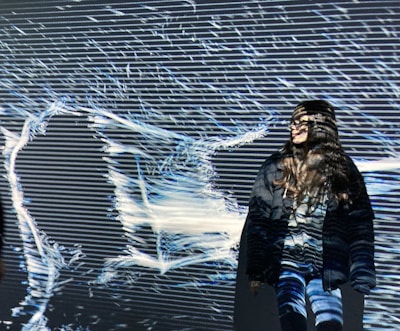 A person stands in front of a light projection that creates abstract patterns and lines, casting shadows on their body. The patterns are predominantly linear and flow across the surface, intertwining with the person's silhouette.