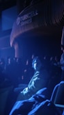 A person sitting in a dimly lit auditorium, illuminated by a bluish spotlight, with other people sitting in the background. The setting appears to be a theater or performance venue with the word 'Turkcell Sahnesi' visible on the wall.