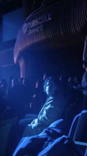 A person sitting in a dimly lit auditorium, illuminated by a bluish spotlight, with other people sitting in the background. The setting appears to be a theater or performance venue with the word 'Turkcell Sahnesi' visible on the wall.
