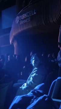 A person sitting in a dimly lit auditorium, illuminated by a bluish spotlight, with other people sitting in the background. The setting appears to be a theater or performance venue with the word 'Turkcell Sahnesi' visible on the wall.