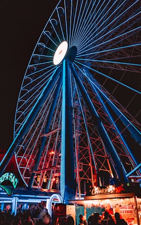 An illuminated Ferris wheel dominates the scene with bright blue lights highlighting its structure. Below, a small food stall with a sign selling churros adds a warm, inviting glow to the atmosphere. The scene captures a nighttime fair setting with people milling around, their silhouettes against the colorful lights.