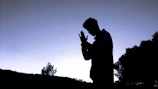 Volunteers praying together outdoors, hands joined, with a backdrop of a sunset sky.