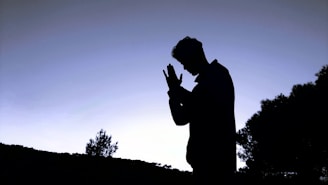 A silhouette of a person with hands clasped in a prayer-like gesture against a twilight sky. Trees and foliage are visible in the background, creating a serene and contemplative atmosphere.