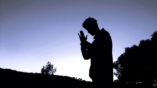 Praying hands silhouetted against a sunrise over a mountain path.