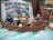 A group of animated characters is depicted in a small boat surrounded by water with foam effects. They appear to be involved in a nautical adventure, with some characters holding oars. The backdrop includes an open comic book with scenes and dialogues related to the adventure.