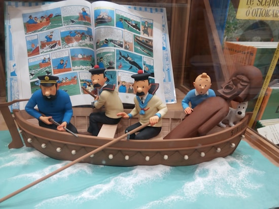 A group of animated characters is depicted in a small boat surrounded by water with foam effects. They appear to be involved in a nautical adventure, with some characters holding oars. The backdrop includes an open comic book with scenes and dialogues related to the adventure.