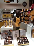 A chocolate shop display featuring a statue of a boy wearing a yellow apron with the Corne Port-Royal logo. Surrounding the statue are various packaged chocolates, including boxes showcasing large assortments and tins with artistic imagery. The shelves are filled with neatly arranged chocolate products, prominently featuring shades of brown and yellow packaging.
