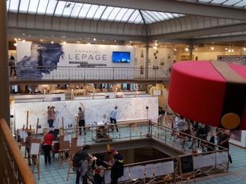 A large art exhibition space with multiple people engaged in live drawing on large canvases. The walls display various artworks, and there's a prominent sign featuring the name Emmanuel Lepage. The environment is well-lit with overhead glass ceiling panels, and the area is bustling with artists and visitors.