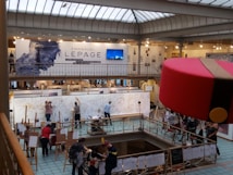 A large art exhibition space with multiple people engaged in live drawing on large canvases. The walls display various artworks, and there's a prominent sign featuring the name Emmanuel Lepage. The environment is well-lit with overhead glass ceiling panels, and the area is bustling with artists and visitors.
