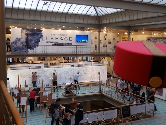 A large art exhibition space with multiple people engaged in live drawing on large canvases. The walls display various artworks, and there's a prominent sign featuring the name Emmanuel Lepage. The environment is well-lit with overhead glass ceiling panels, and the area is bustling with artists and visitors.