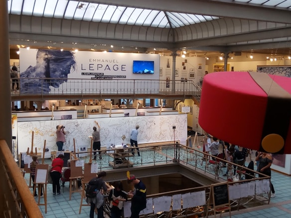 A large art exhibition space with multiple people engaged in live drawing on large canvases. The walls display various artworks, and there's a prominent sign featuring the name Emmanuel Lepage. The environment is well-lit with overhead glass ceiling panels, and the area is bustling with artists and visitors.