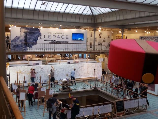 A large art exhibition space with multiple people engaged in live drawing on large canvases. The walls display various artworks, and there's a prominent sign featuring the name Emmanuel Lepage. The environment is well-lit with overhead glass ceiling panels, and the area is bustling with artists and visitors.